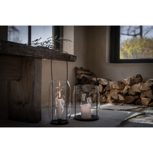 Lyhdyssä on musta metallinen kynttilänalunen ja kahva sekä lasisylinteri jonka sisälle kynttilä laitetaan.