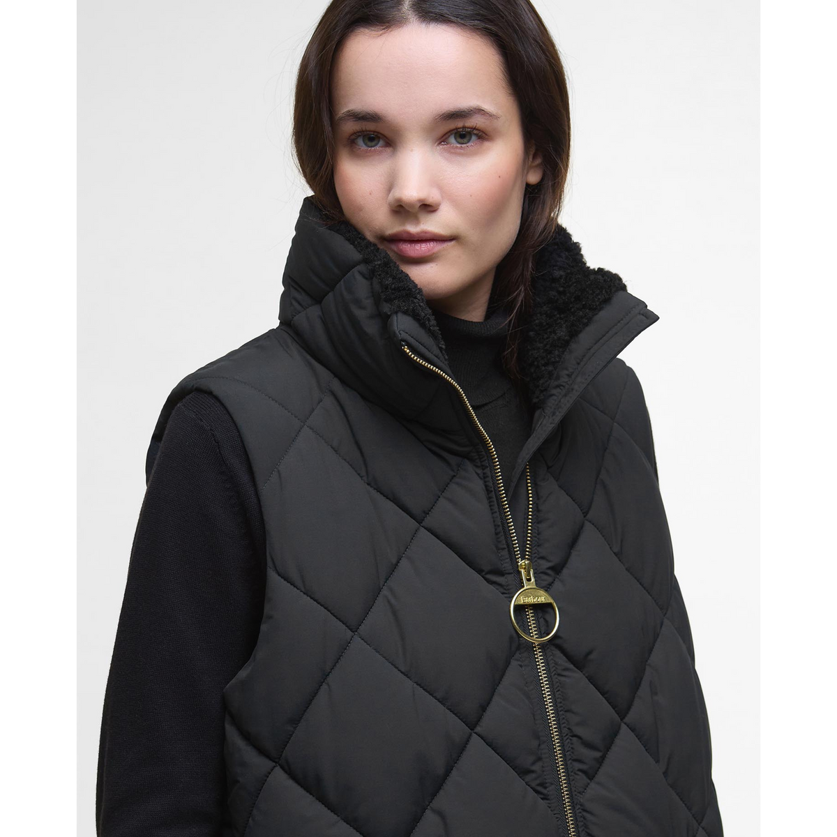 Kuvassa naisella on musta Barbour Bernice Puffer toppaliivi jossa on pehmeä teddyvuorattu kaulus ja näyttävä kullanvärinen vetoketju.