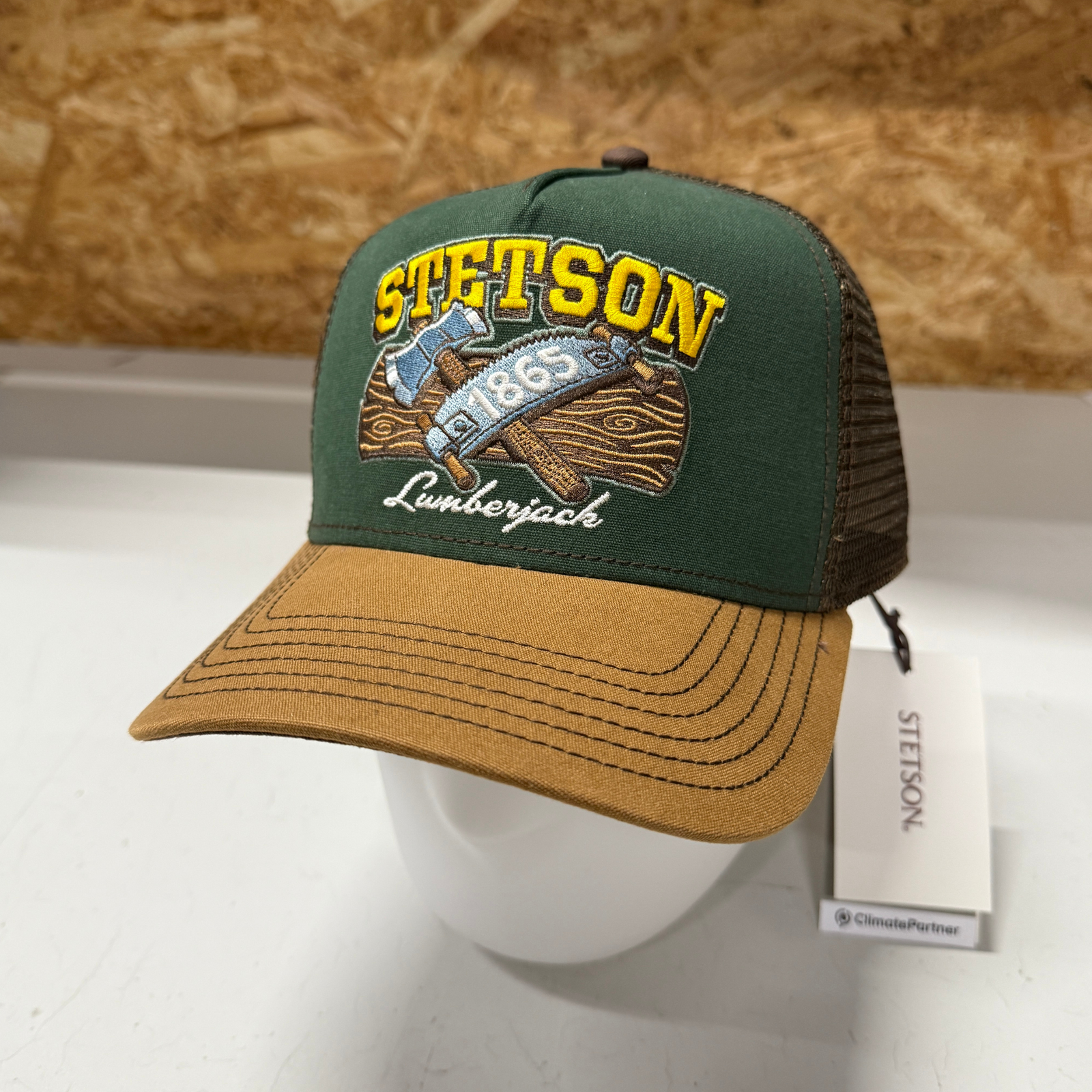 Kuvassa Stetson Lumberjack Trucker Cap jossa ruskea verkko-osa takana, ruskea lippa ja vihreä etuosa. Etuosassa on kelta-ruskea Stetson Lumberjack -brodeeraus.