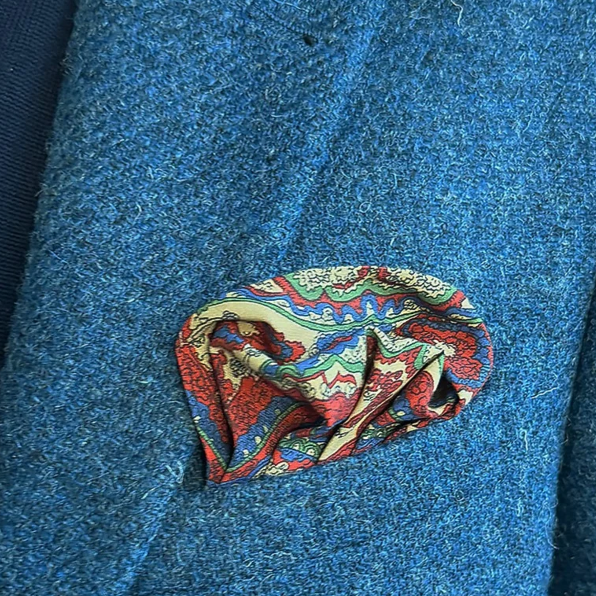 Wellington of Bilmore monivärinen silkkitaskuhuivi sinisen tweedbleiserin rintataskussa, pyöreästi laskostettuna.